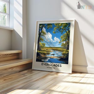 Op de afbeelding: Een ingelijste print van een schilderachtig landschap met een rivier die door een weelderig groen bos stroomt met een blauwe lucht en witte wolken. De tekst "EVERGLADES NATIONAL PARK" is onder de afbeelding gedrukt.