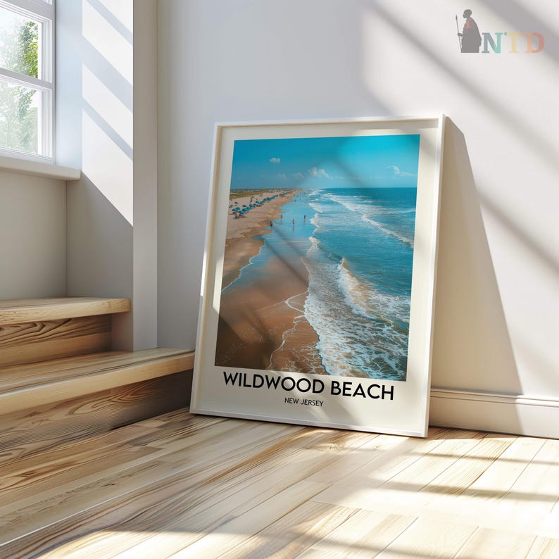 May include: A framed print of Wildwood Beach, New Jersey, showing a beach scene with a blue sky and ocean. The print is in a white frame and leans against a white wall on a wooden floor.
