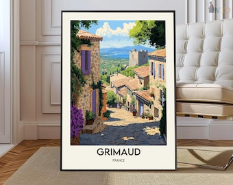 May include: Framed art print of Grimaud, France. The artwork depicts a cobblestone street lined with stone buildings, purple shutters, and flowering plants. The background shows a scenic view of the sea and sky. The print is in a black frame.