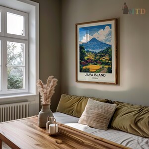 May include: A framed art print titled "JAVA ISLAND INDONESIA" depicts a scenic landscape with a mountain, fields, and village. The print is in a wooden frame and hangs on a neutral-colored wall above a sofa with olive-green cushions and a decorative pillow.