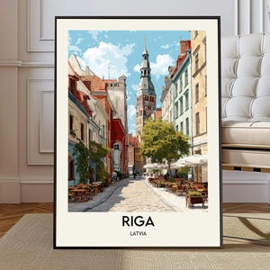 May include: A framed art print of a European street scene in Riga, Latvia. The image features colorful buildings, a tall clock tower, and outdoor seating. The sky is blue with fluffy clouds. The word "RIGA" is printed at the bottom.