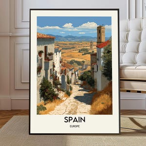 May include: A framed art print depicting a sunlit street scene in Spain. The artwork features white buildings with red-tiled roofs, a cobblestone street, and a distant landscape under a blue sky. The word "SPAIN" and "EUROPE" are printed at the bottom.