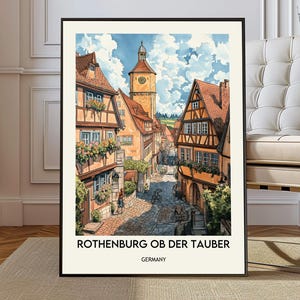 May include: A framed watercolour print of Rothenburg ob der Tauber, Germany. The artwork shows a cobbled street with colourful buildings, a clock tower, and a blue sky with clouds. The text "ROTHENBURG OB DER TAUBER GERMANY" is at the bottom.