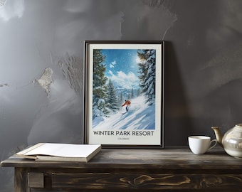May include: Framed poster showing a skier on a snowy slope at Winter Park Resort, Colorado. The artwork depicts a sunny day with snow-covered trees and a blue sky. The poster is displayed on a dark wooden table.