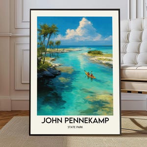 Op de afbeelding: Een ingelijste kunstdruk van John Pennekamp State Park. Het kunstwerk toont een turquoise waterweg met een kajak, palmbomen en een helderblauwe lucht met witte wolken. De tekst "JOHN PENNEKAMP STATE PARK" staat onderaan.