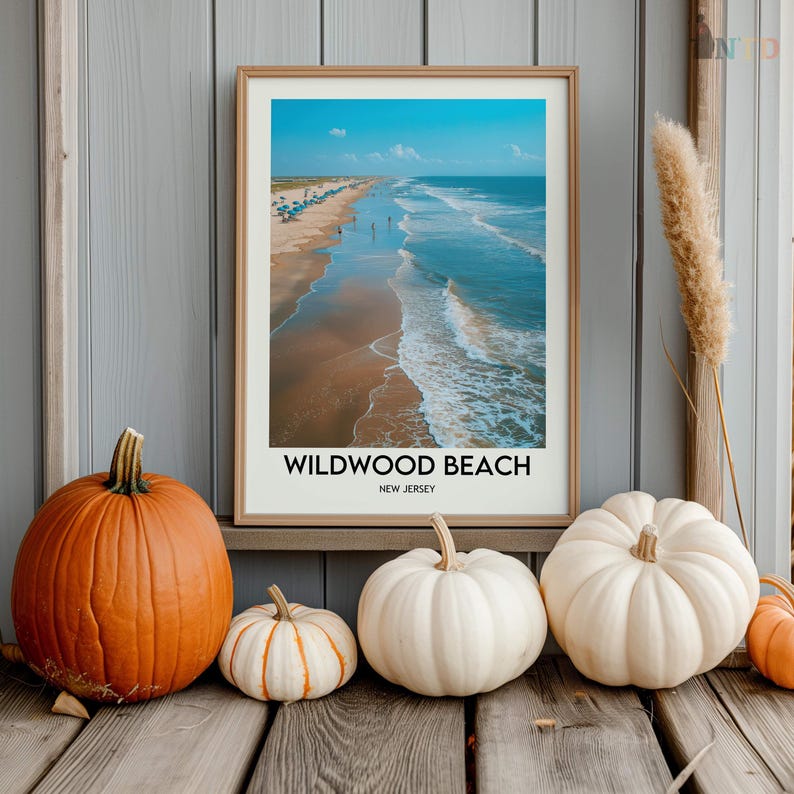 May include: A framed print of Wildwood Beach, New Jersey, with a blue sky and ocean. The print is displayed with pumpkins in orange and white, creating a seasonal autumn theme. The pumpkins are on a wooden surface.