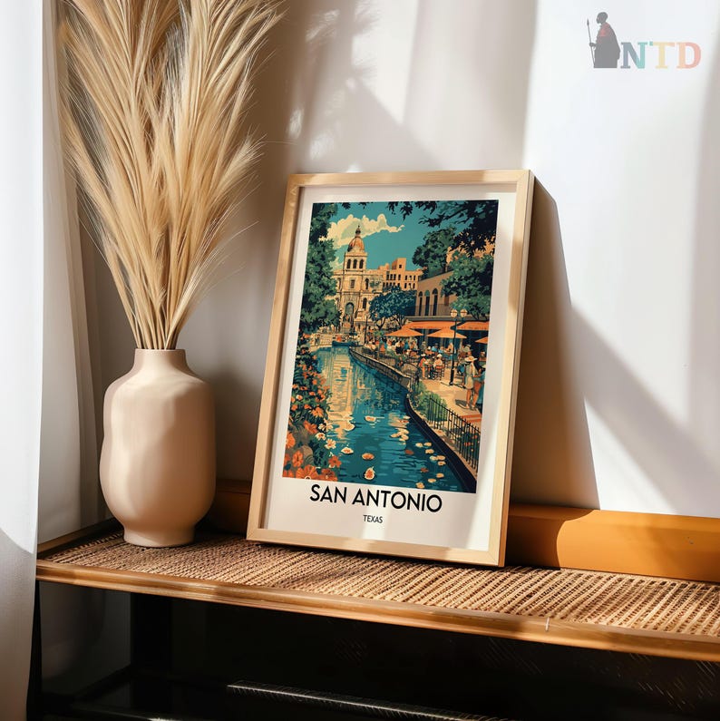 May include: A framed print of a colorful painting depicting a canal in San Antonio, Texas. The painting features a blue canal with buildings and trees on either side. The canal is filled with water and there are people walking along the banks. The print is framed in a wooden frame.