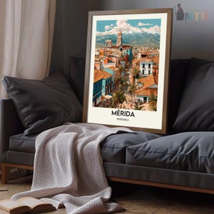 K&ouml;nnte beinhalten: Ein gerahmter Kunstdruck mit einer farbenfrohen Illustration von M&eacute;rida, Venezuela. Das Kunstwerk zeigt eine Stadtlandschaft mit roten Ziegeld&auml;chern, blauen Geb&auml;uden und einer Bergkette im Hintergrund. Der Druck wird auf einem grauen Sofa mit einer Decke pr&auml;sentiert.
