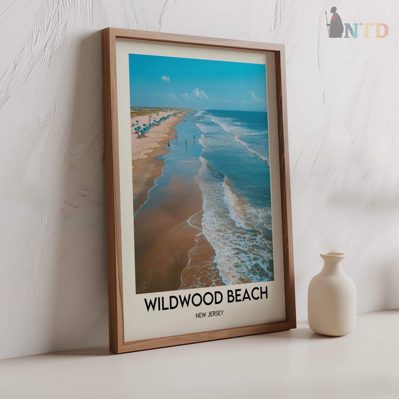 May include: A framed print of Wildwood Beach, New Jersey, showing a beach scene with a blue sky, ocean waves, and sandy shore. The print is in a light brown wooden frame. A small, off-white vase is to the right.