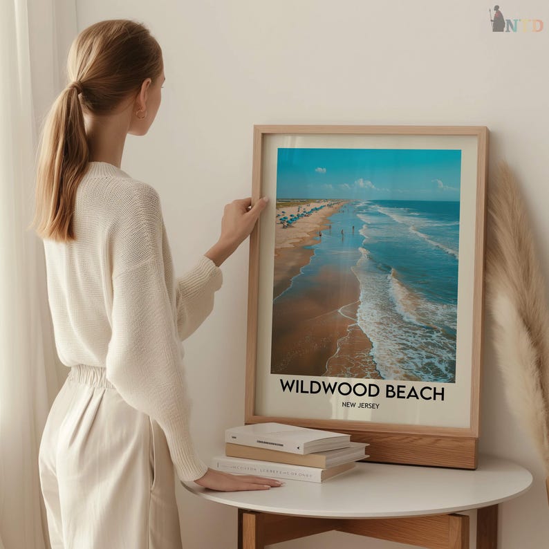 May include: A framed print of Wildwood Beach, New Jersey, depicting a coastal scene. The artwork shows a sandy beach, ocean waves, and a clear blue sky. The print is presented in a light-coloured wooden frame.