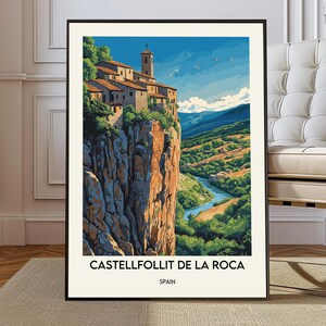 May include: A framed art print of Castellfollit de la Roca, Spain. The image shows a village on a cliff, with a church tower and buildings with red tile roofs. The landscape includes a river, green hills, and a blue sky.