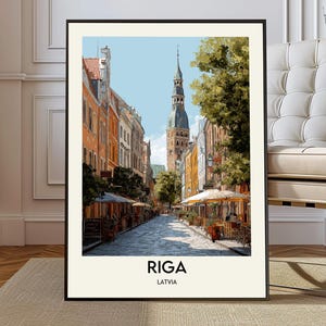 Op de afbeelding: Ingelijste kunstdruk met een aquarel schilderij van Riga, Letland. Het kunstwerk toont een geplaveide straat met kleurrijke gebouwen, cafés en een hoge kerktoren onder een blauwe hemel. De woorden "RIGA" en "LATVIA" staan onderaan afgedrukt.