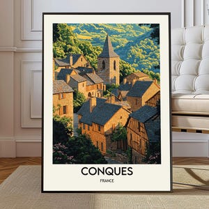 May include: A framed art print featuring a picturesque village scene. The image showcases stone buildings with dark rooftops, a tall church steeple, and lush green hills. The word "CONQUES" and "FRANCE" are printed at the bottom.