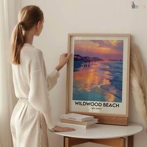 K&ouml;nnte beinhalten: Ein gerahmter Druck von Wildwood Beach, New Jersey, mit einem Sonnenuntergang &uuml;ber dem Meer. Das Kunstwerk zeigt eine Strandszene mit Personen, einem Pier und dem Text "Wildwood Beach, New Jersey". Der Rahmen ist aus hellem Holz.
