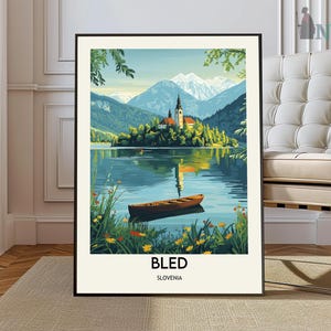 Könnte beinhalten: Ein gerahmter Kunstdruck vom Bleder See, Slowenien. Das Kunstwerk zeigt eine ruhige Seeszene mit einer Kirche auf einer Insel, Bergen im Hintergrund und einem Holzboot. Der Druck hat einen schwarzen Rahmen und das Wort "BLED" ist unten aufgedruckt.