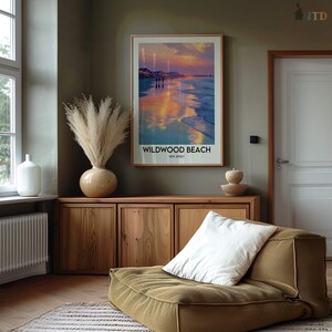 K&ouml;nnte beinhalten: Ein gerahmtes Poster mit den Worten "Wildwood Beach, New Jersey" zeigt eine Strandszene mit einem farbenfrohen Sonnenuntergang. Das Poster h&auml;ngt &uuml;ber einem Holzschrank, neben einer Vase mit hohen, getrockneten Pflanzen. Ein gr&uuml;nes Bodenkissen mit einem wei&szlig;en Kissen befindet sich im Vordergrund.