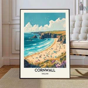 May include: A framed art print of Cornwall, England, featuring a beach scene with turquoise water, a sandy shore, and cliffs. The artwork has a vintage travel poster style, with people and umbrellas on the beach under a blue sky with clouds.