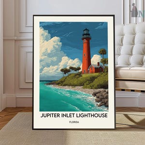 May include: A framed art print of the Jupiter Inlet Lighthouse in Florida. The artwork depicts a tall, red lighthouse next to a small red building, set against a blue sky with fluffy white clouds. The turquoise ocean meets a sandy shore.