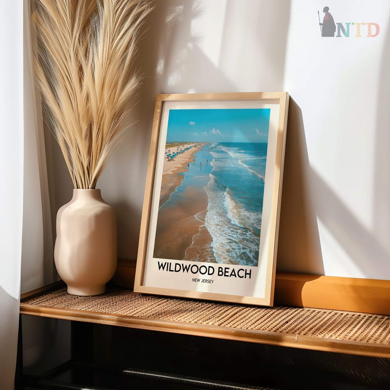 May include: A framed print of Wildwood Beach, New Jersey, in a light wood frame. The image depicts a beach with blue ocean water, waves, and people. A beige vase with tall, dried grass is positioned to the left of the framed print.