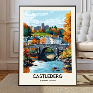 Może przedstawiać: Oprawiony wydruk artystyczny przedstawiający Castlederg w Irlandii Północnej, z kamiennym mostem nad rzeką, kolorowymi domami i zamkiem w tle. Grafika ma żywą paletę barw z jesiennymi liśćmi.