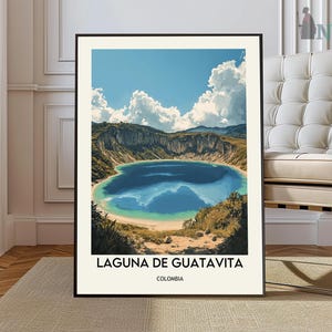 May include: A framed art print featuring a scenic view of Laguna de Guatavita in Colombia. The artwork depicts a blue lake surrounded by green hills under a bright blue sky with fluffy white clouds. The text "LAGUNA DE GUATAVITA COLOMBIA" is at the bottom.
