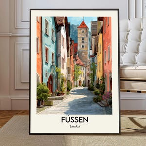 May include: A framed print of a European street scene. The artwork shows a narrow cobbled street flanked by colourful buildings in shades of orange, blue, and yellow. A tall clock tower is visible in the distance. The text "FÜSSEN" and "BAVARIA" are at the bottom.