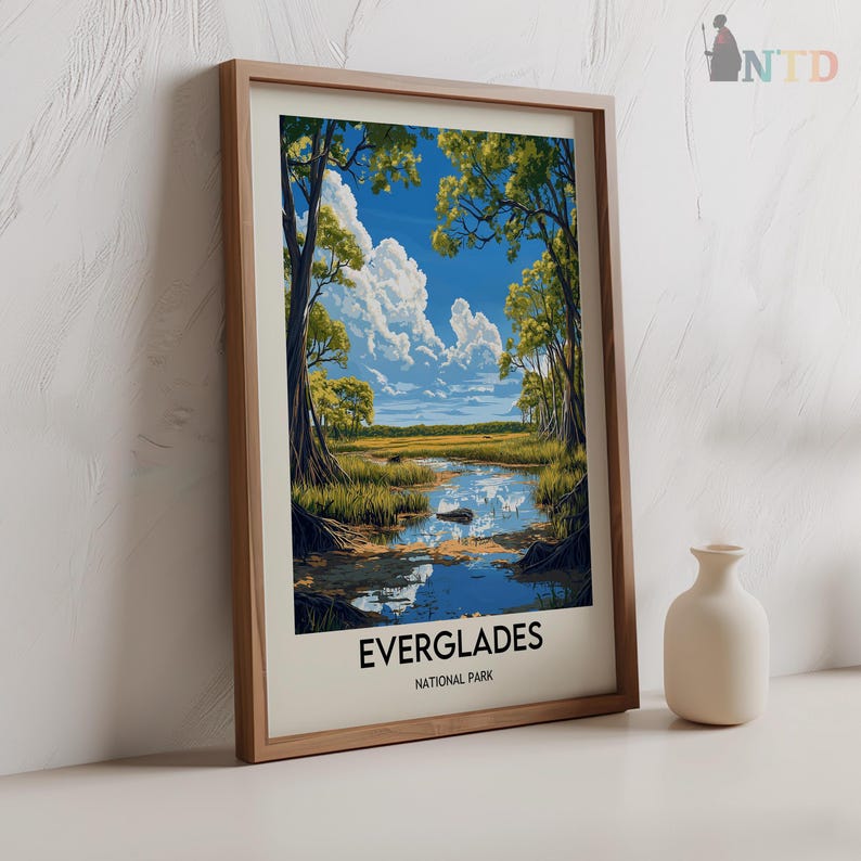Op de afbeelding: Een ingelijste print van een landschapssc&egrave;ne met een rivier die door een grasachtig gebied stroomt. De rivier wordt omringd door bomen en de lucht is blauw met witte wolken. De print heeft de tekst "Everglades National Park" onderaan.