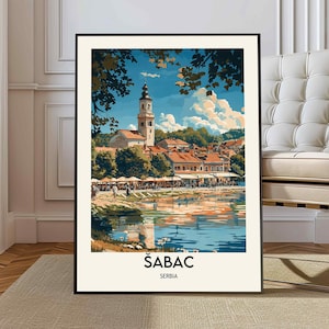 Può includere: Una stampa artistica incorniciata di Šabac, Serbia, con un'illustrazione dettagliata di una città sul fiume con un campanile. L'opera d'arte presenta una vivace tavolozza di colori con cieli blu, tetti rossi e riflessi nell'acqua.