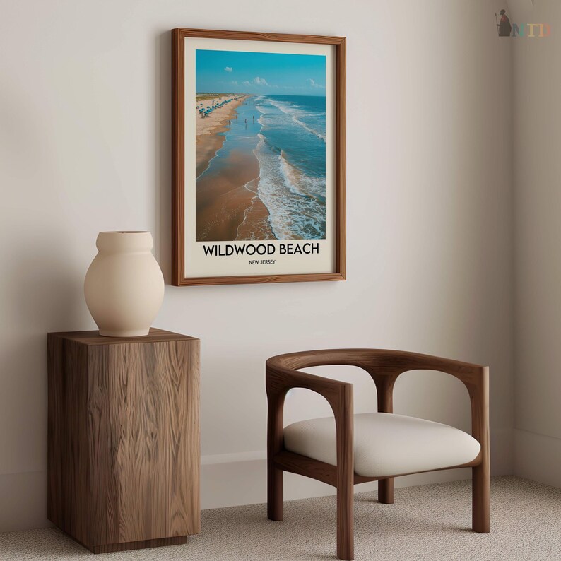 May include: A framed print of Wildwood Beach, New Jersey, with a brown wooden frame. The print shows a beach scene with blue water, a sandy shore, and people. A beige vase sits on a wooden side table, and a wooden chair with a white cushion is nearby.