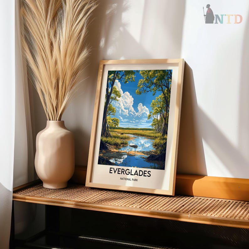 Op de afbeelding: Een ingelijste print van een landschapssc&egrave;ne met een blauwe lucht, witte wolken en groene bomen die worden weerspiegeld in een stil waterlichaam. De print is getiteld "Everglades National Park".