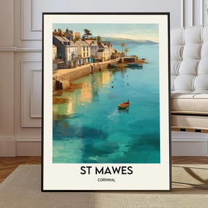 Op de afbeelding: Een ingelijste kunstdruk met een kustgezicht van St. Mawes, Cornwall, met een rij huizen langs een stenen havenmuur, een klein bootje in turquoise water en de tekst ST MAWES CORNWAL in zwarte letters onderaan de crèmekleurige rand.