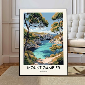 Puede incluir: Una impresión artística enmarcada que muestra una vista de Mount Gambier, Australia. La obra de arte representa una bahía turquesa rodeada de acantilados, árboles y un cielo azul. El texto "MOUNT GAMBIER AUSTRALIA" está impreso en la parte inferior.