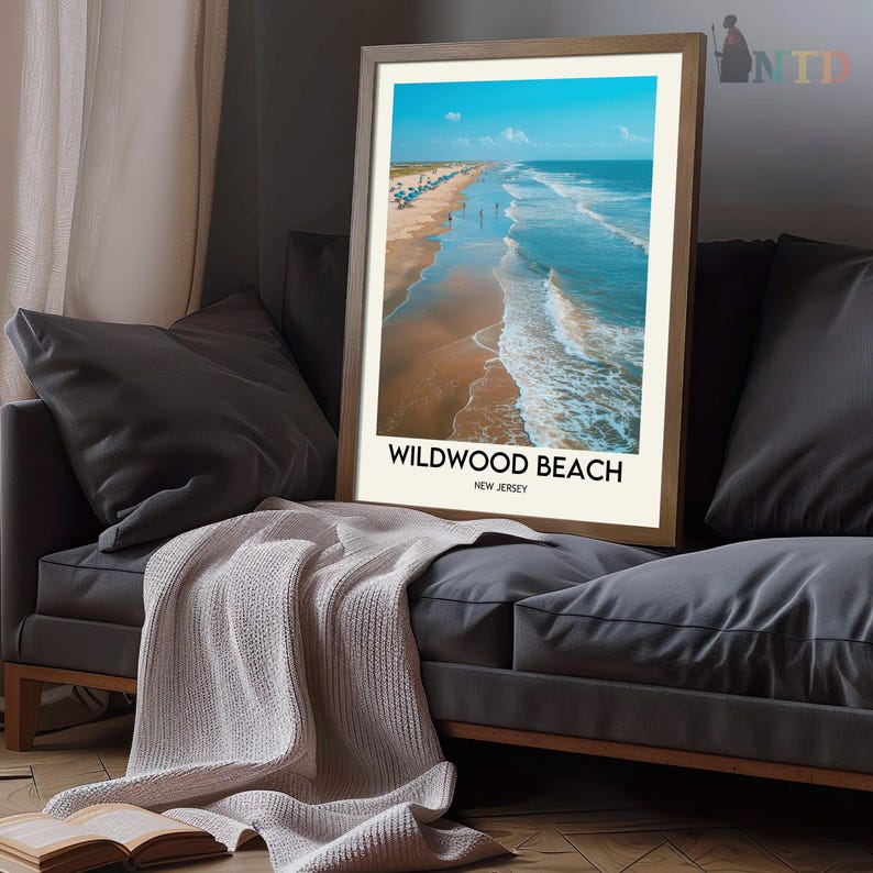 May include: A framed print of Wildwood Beach, New Jersey, showing a beach scene with blue ocean waves and a clear sky. The print is in a wooden frame and rests on a grey sofa with pillows and a light-coloured blanket. The text "Wildwood Beach, New Jersey" is displayed.