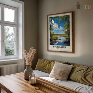 Op de afbeelding: Een ingelijste vintage poster met een schilderachtig uitzicht op het Everglades National Park. De poster toont een blauwe lucht met witte wolken, groene bomen en een kalm wateroppervlak dat de lucht weerspiegelt. De tekst op de poster luidt "Everglades National Park".