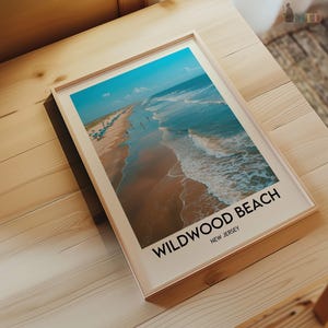 May include: A framed print depicting a beach scene with turquoise water and a sandy shore. The print includes the text "Wildwood Beach, New Jersey" in a bold, black font. The frame is a light wood colour.