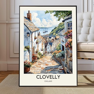 May include: Watercolor print of Clovelly, England, featuring a cobblestone street winding through white buildings with flower boxes. The artwork depicts a coastal village with a view of the sea and sailboats under a blue sky. The print is framed in black.