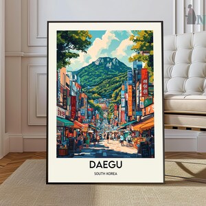May include: A framed art print of Daegu, South Korea, depicting a bustling street scene with shops, restaurants, and pedestrians. The artwork features vibrant colors, a mountain backdrop, and the city's name in bold text.
