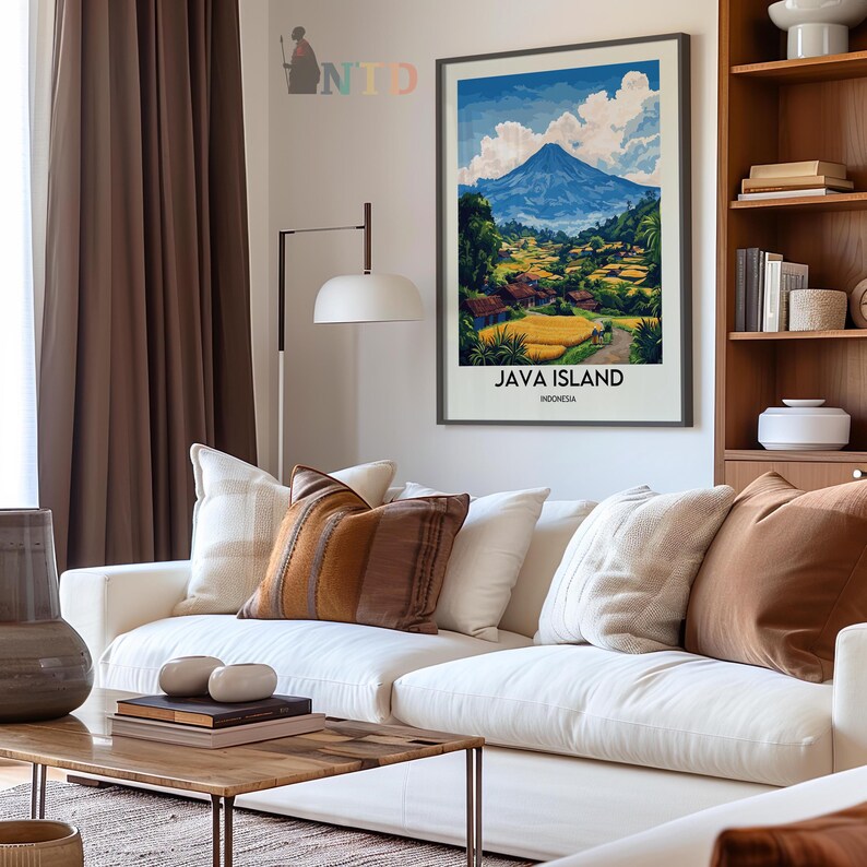 May include: A framed art print of Java Island, Indonesia, featuring a mountain landscape with a blue sky and white clouds. The print is displayed above a white sofa with decorative pillows in shades of brown and cream. A wooden coffee table and a bookshelf are also visible.