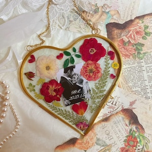 May include: A heart-shaped, gold-framed photo holder with a black and white photo of a couple. The frame is filled with dried red and pink flowers and greenery. The photo has the initials "R & L" and the date "2025 16". A gold chain is attached for hanging.
