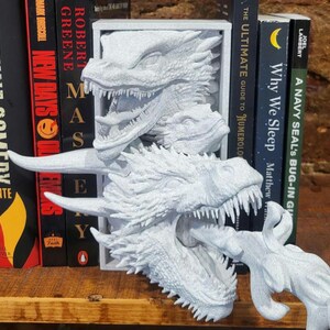 Pu&ograve; includere: Un set di fermalibri a tema drago bianco. Il set include un fermalibri a forma di testa di drago e un elemento di fuoco di drago separato. I fermalibri sono posizionati davanti a una fila di libri.