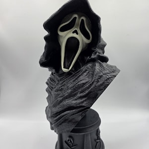 May include: A black bust of a hooded figure with a pale, elongated mask featuring large, dark eye sockets and an open, screaming mouth. The figure wears a textured, draped robe and stands on a cylindrical base decorated with skull and crossbones.
