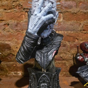 Op de afbeelding: Een lichtblauw borstbeeld van een figuur met een raster van zilveren spelden in het hoofd en gezicht. De figuur houdt één hand voor de ogen en neus. Het draagt een zwart, getextureerd kledingstuk op een zwarte voet met een sculptuur.