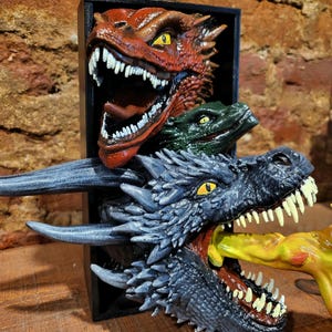 Pu&ograve; includere: Scultura di drago tridimensionale con tre teste di drago rosse, verdi e grigie. La testa di drago grigia &egrave; in primo piano, con un oggetto giallo in bocca. La scultura &egrave; ambientata su uno sfondo nero.
