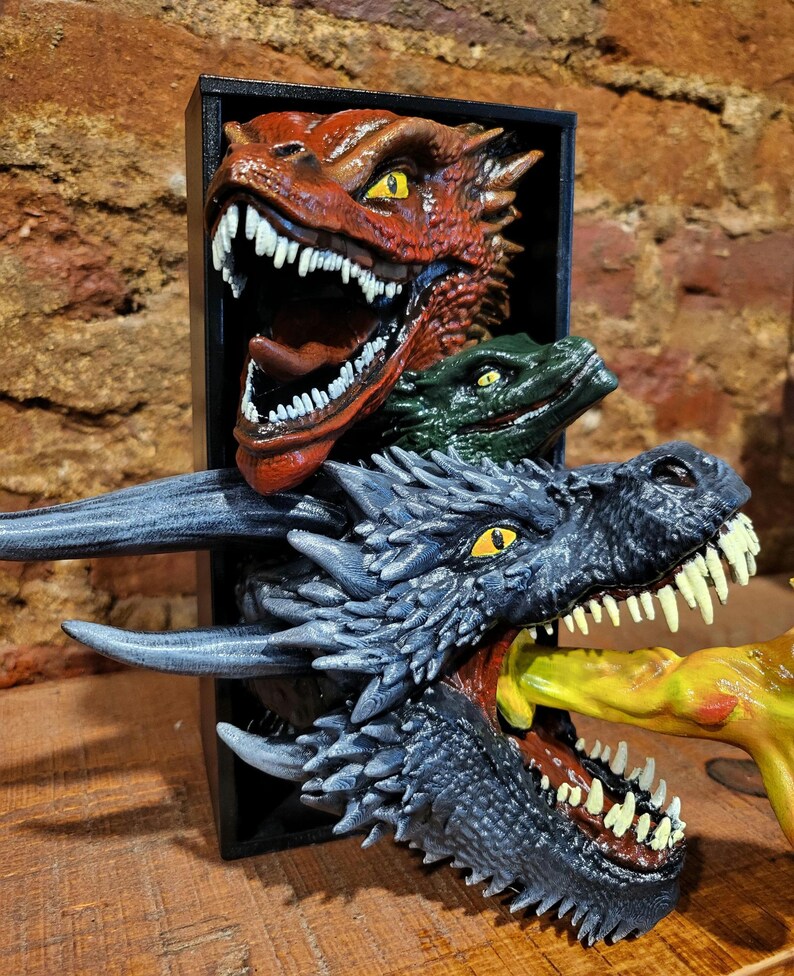 Pu&ograve; includere: Scultura di drago tridimensionale con tre teste di drago rosse, verdi e grigie. La testa di drago grigia &egrave; in primo piano, con un oggetto giallo in bocca. La scultura &egrave; ambientata su uno sfondo nero.
