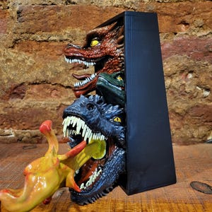 Pu&ograve; includere: Una scultura rettangolare nera con tre teste di drago rosse, verdi e blu, che emergono da una scatola scura. Una creatura gialla e rossa viene divorata dal drago blu. La scultura &egrave; su una superficie di legno.