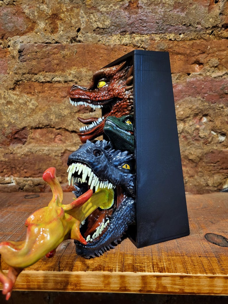 Pu&ograve; includere: Una scultura rettangolare nera con tre teste di drago rosse, verdi e blu, che emergono da una scatola scura. Una creatura gialla e rossa viene divorata dal drago blu. La scultura &egrave; su una superficie di legno.