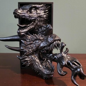 Pu&ograve; includere: Scultura di drago nero con tre teste di drago che emergono da una cornice rettangolare. I draghi hanno squame dettagliate, denti affilati e un effetto di soffio di fuoco. La scultura &egrave; su una superficie di legno.
