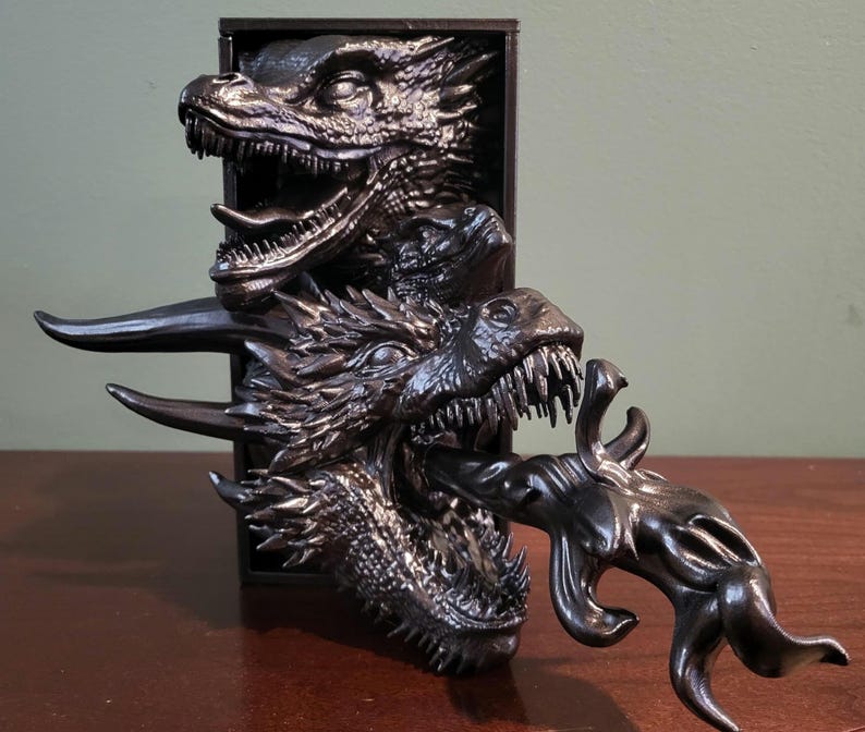 Pu&ograve; includere: Scultura di drago nero con tre teste di drago che emergono da una cornice rettangolare. I draghi hanno squame dettagliate, denti affilati e un effetto di soffio di fuoco. La scultura &egrave; su una superficie di legno.
