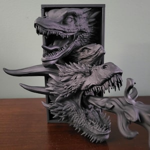Pu&ograve; includere: Scultura di drago grigio scuro con tre teste di drago che emergono da una cornice rettangolare nera. La testa inferiore del drago &egrave; aperta, con un getto di fuoco. La scultura &egrave; su una superficie marrone.