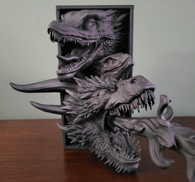 Pu&ograve; includere: Scultura di drago grigio scuro con tre teste di drago che emergono da una cornice rettangolare nera. La testa inferiore del drago &egrave; aperta, con un getto di fuoco. La scultura &egrave; su una superficie marrone.
